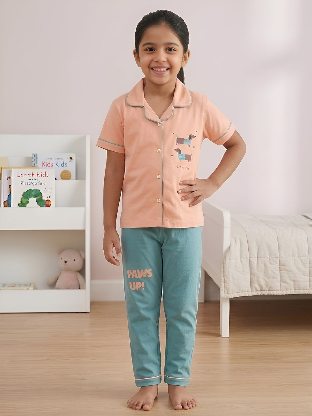 Child wearing a peach shirt and teal pants with 'PAWS UP!' text in a bedroom setting.